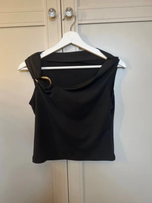 Svart oneshoulder topp med guldring - Säljer en svart oneshoulder topp med snygg guldfärgad ringdetalj på axeln. Toppen är ärmlös och har en cool, asymmetrisk design som ger en trendig look. Perfekt för dig som vill sticka ut lite extra.