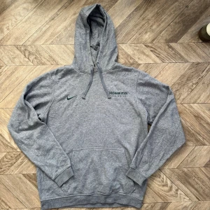 Grå Michigan State hoodie - Säljer en grå hoodie med huva och magficka. På bröstet står det 'Michigan State Field Hockey' och den har en liten Nike-logga i grönt. Hoodien har ribbade muddar vid ärmar och nederkant samt dragsko i huvan. Perfekt för chill dagar eller träning. Den har ett knappt synlig flöck i mitten av framsidan.