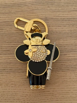  Vuitton samuraj nyckelring - Unik nyckelring från  Vuitton i form av en samurajfigur. Guld- och svartfärgad med gnistrande stenar och detaljer som ett litet svärd. Tillverkad i metall och emalj med exklusiv finish och LV-logga på flera ställen. Perfekt accessoar för att piffa upp väskan eller nyckelknippan.