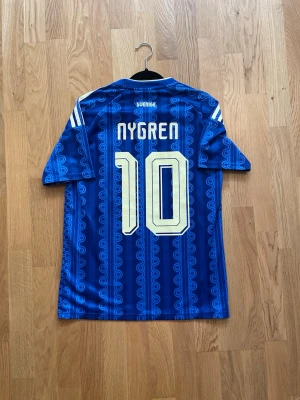 Nygren #10 - Säljer en blå fotbollströja med Sveriges landslagsemblem och nummer 10 på både fram- och baksidan. Tröjan har vita detaljer, tre ränder på axlarna och namnet Nygren på ryggen. Tillverkad i ett lätt och ventilerande material, perfekt för träning eller match.