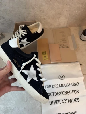Golden goose  - Snygga svarta sneakers från Golden Goose med vit stjärndetalj på sidan och vit häl. Skorna har vita snören och en robust, något smutsad sula för en cool vintage-look. Ovandelen är i mocka och detaljerna i läder. Perfekta för dig som gillar streetstyle.