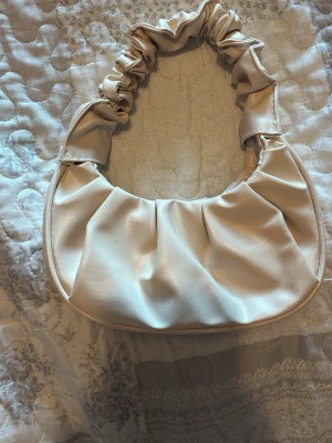 Krämvit axelväska med rynkad rem - Trendig handväska i krämvitt konstläder med en snygg rynkad axelrem. Väskan har en mjuk, rundad form och veckad design framtill som ger en cool och modern vibe. Perfekt accessoar för att lyfta din outfit.