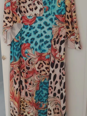 Långklänning med leopardmönster - Unik långklänning med leopardmönster i blått, beige, svart och röda toner. Klänningen har vida ärmar och ett flowy fall, perfekt för dig som vill sticka ut. Materialet känns lätt. Oanvänd. 
