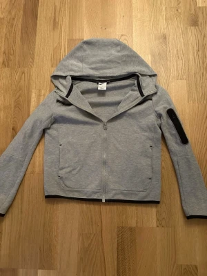 Nike Tech - Snygg grå hoodie från Nike med hel dragkedja framtill och svarta detaljer. Tröjan har huva, två sidofickor och en extra ficka på ena ärmen. Tillverkad i mjukt sweatshirtmaterial, perfekt för en avslappnad streetwear-look. Bra och värd pris!