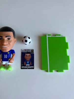 Zuru FIFA Ballers Fotbollsfigur Kylian Mbappé set - Säljer ett samlarset med en Kylian Mbappé-figur i blått matchställ, en liten svartvit fotboll, ett grönt plastunderlag som ser ut som en fotbollsplan och ett samlarkort. Perfekt för dig som gillar fotboll och vill ha något kul till hyllan eller skrivbordet.