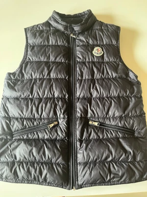 Svart dunväst från Moncler - Snygg svart dunväst från Moncler med hög krage och två dragkedjefickor framtill. Västen har Moncler-logga på bröstet och quiltad design som ger en modern look. Perfekt att slänga över en hoodie eller tröja för extra värme.