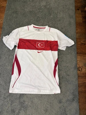Turkiet fotbollströja Nike vit/röd - Säljer en vit fotbollströja från Nike med Turkiets flagga på bröstet och röda detaljer. Tröjan har rund hals, korta ärmar och tryck på båda ärmarna. Perfekt för dig som vill stötta Turkiets landslag.