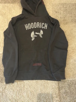 Svart hoodie från Hoodrich med tryck - Svart hoodie från Hoodrich med stor vit text på ryggen och två vita fåglar tryckta under. Har en klassisk huva och magficka samt röd textdetalj vid fickan. Perfekt för dig som gillar streetwear och vill sticka ut. XL i barnstorlek