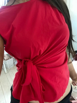Röd omlottopp med knyt i midjan - Snygg röd omlottopp med knyt i midjan och ärmlös design. Toppen har en enkel, stilren look och är tillverkad i ett mjukt tyg som känns skönt mot huden. Perfekt för dig som vill ha en färgstark och trendig topp till din outfit.