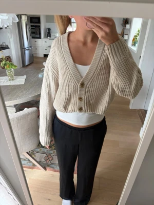 Beige cropped kofta från Carin Wester  - Beige, ribbstickad cropped kofta från C.W. med djup V-ringning och tre bruna knappar framtill. Koftan har långa ärmar och är gjord i ett mjukt, tjockt material. 