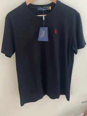 Svart t-shirt från Polo Ralph Lauren - Klassisk svart t-shirt från Polo Ralph Lauren med röd broderad logga på bröstet. T-shirten har rund halsringning och korta ärmar. Tillverkad i mjuk bomull för en skön känsla och enkel stil.