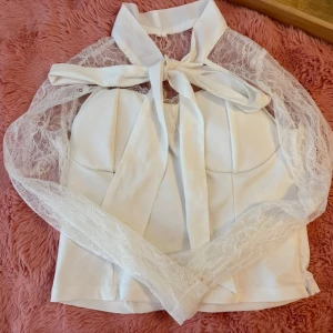 Vit blus med spetsärmar  - Supersöt vit blus med transparenta spetsärmar och stor rosett i halsen. Toppen har en croppad passform och ärmlösa axlar under spetsen. Perfekt för dig som vill sticka ut med en romantisk och trendig look.
