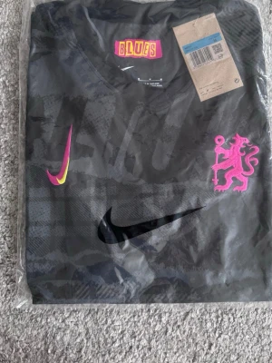 Svart Nike fotbollströja med rosa detaljer chealsea - Snygg svart fotbollströja från Nike med grafiskt mönster och rosa detaljer på ärmar och logga. Tröjan har rund hals, korta ärmar och är tillverkad i Dri-FIT material som andas. Perfekt för träning eller match! Chealsea