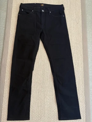 Svarta jeans från Lee W32 L32 - Snygga svarta jeans med klassisk femficksdesign och raka ben. Jeansen har silverfärgade nitar och knappdetaljer samt bälteshällor. Materialet ser ut att vara en stretchig bomullsblandning för extra komfort. Helt oanvända 
