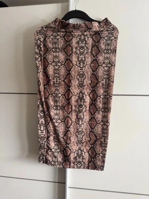 Brun kjol med ormmönster - Säljer en snygg, midilång kjol med ormmönster i bruna och beige toner. Kjolen har en rak passform och är tillverkad i ett mjukt, stretchigt material som sitter bekvämt. Perfekt för dig som vill sticka ut med ett trendigt mönster.
