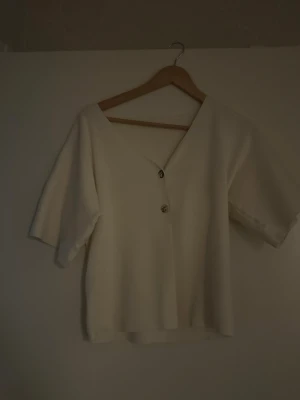 Vit v-ringad topp med knappar - Stilren vit topp med v-ringning och två dekorativa knappar framtill. Toppen har korta, vida ärmar och rak passform. Perfekt att matcha med jeans eller kjol för en enkel men snygg look.
