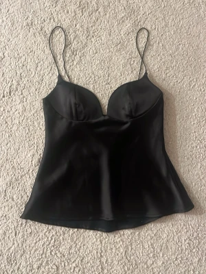 Svart satinlinnetopp från Zara - Snygg svart linnetopp från Zara i satinliknande material. Toppen har tunna axelband och formade kupor som ger en feminin siluett. Perfekt att styla med jeans eller kjol för en trendig look.