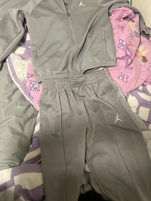 Grått Jordan loungewear set - Snyggt grått loungewear set från Jordan med dragkedja på tröjan och elastisk midja på byxorna. Setet har vita Jordan-loggor på både tröja och byxor samt diskreta fickor. Perfekt för chill dagar hemma eller när du vill vara bekväm men ändå se fresh ut.