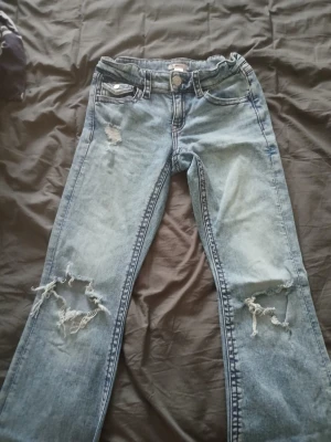 Ljusblå slitna jeans med hål - Säljer ett par ljusblå jeans med slitningar och stora hål vid knäna. Jeansen har klassisk femficksdesign och knappgylf. Baktill finns fickor med lock och knapp. Perfekta för en avslappnad och trendig look.