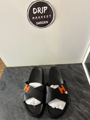 sandaler med orange H-detalj - Säljer ett par stilrena svarta sandaler i storlek 43 i läder. med korsade remmar i läder och en tydlig orange H-detalj på varje sandal. Perfekta för dig som vill ha en clean look med en twist. Sula i svart och öppen tå för skön sommarkänsla.