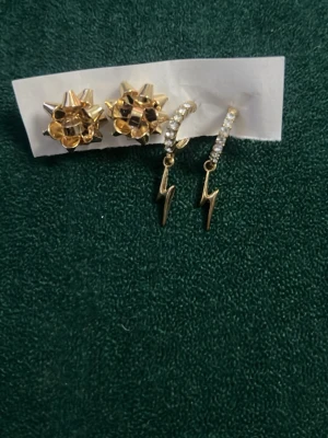 Två par örhängen i guld och pärlor - Säljer två par helt örhängen: ett par i guldton med rosettform och ett par med blixtformade hängen. Det andra paret har små vita pärlor på bågen och guldiga blixtar som hänger ner. Perfekt för dig som gillar unika accessoarer. Och i väldigt bra skick. Har aldrig använt de. 