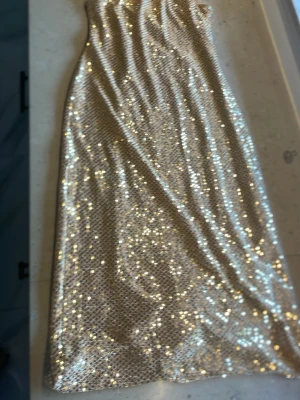 Glittrig guldig axelbandslös klänning - Säljer en glittrig axelbandslös klänning i guld med paljetter. Klänningen är kort och har en mjuk beige insida för extra komfort. Perfekt för dig som vill sticka ut och glänsa på nästa fest.