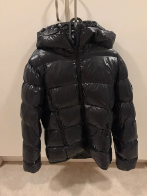 Svart glansig dunjacka från Everest - Snygg svart dunjacka från Everest med glansig finish, huva och två dragkedjeförsedda fickor framtill. Jackan har en puffig look och är perfekt för kalla vinterdagar. Jackan är i strl 40. Den är väldigt varm och har verkligen varit perfekt till vintern. 