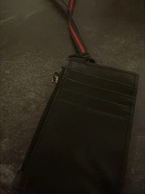 Svart plånbok med orm och rem - Snygg svart plånbok i avlång modell med flera kortfack och dragkedja. Ena sidan har ett tryck av en röd, vit och svart orm. Plånboken har en avtagbar rem i svart och rött som gör den enkel att bära runt halsen eller handleden.