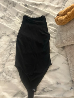 Svart ärmlös body med hög hals - Säljer en enkel och stilren svart body med hög hals och ärmlös design. Bodyn har tryckknappar i grenen för enkel på- och avklädning. Perfekt att matcha med jeans eller kjol för en clean look.