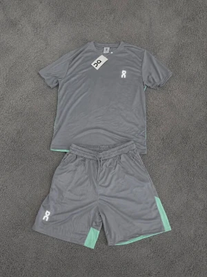 On Running träningsset - Ett snyggt och bekvämt set med t-shirt och shorts från On Running, perfekt för träning eller fritid. Båda plaggen är i grått med detaljer i en ljusgrön färg. T-shirten har en rund hals och korta ärmar, medan shortsen har en elastisk midja med dragsko och en innerbyxa. Båda plaggen har On Runnings logotyp. Specifikationer:  Märke: On Running  Storlek: S  Färg: Grå/ljusgrön  Material: 100% polyester  Skick: Helt nytt med lappar kvar Detta set är perfekt för dig som söker löp kläder