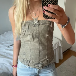 Grå jeanskorsett från Zara - Snygg grå jeanskorsett från Zara med knappar framtill och dekorativa sömmar. Grå men har en lite olivgrön ton enligt mig. Den har en unik design med bältesdetaljer och är perfekt för att ge en edgy touch till din outfit. Passar bra till både jeans och kjol. 