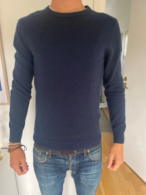 Mörkblå stickad tröja herr - Mörkblå stickad tröja med rund hals och långa ärmar. Tröjan har en enkel, stilren design utan synliga loggor eller mönster. Materialet ser mjukt och lite stretchigt ut, perfekt för lager på lager under kyligare dagar. Model 185, 71kg bär S. Hör av er vid frågor