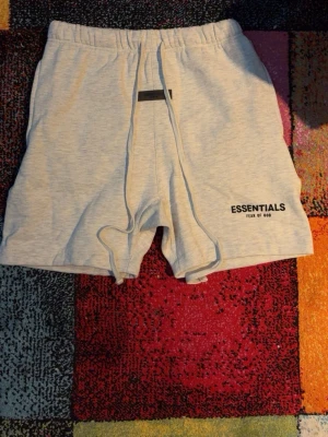 Grå Essentials Fear of God shorts - Snygga ljusgrå shorts från Essentials Fear of God med elastisk midja och snörning framtill. Tillverkade i mjuk bomull för maximal komfort. Loggan är tryckt på vänster ben. Perfekta för chill dagar eller streetwear-stil.