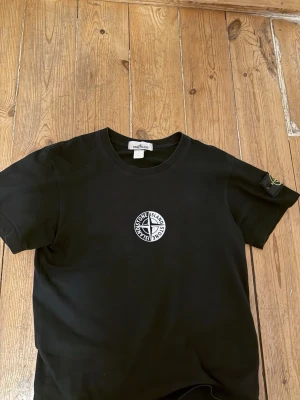 Svart Stone Island t-shirt - Säljer en svart t-shirt från Stone Island med den klassiska loggan tryckt på bröstet och en patch på vänster ärm. Qr code fungerar utmärkt 