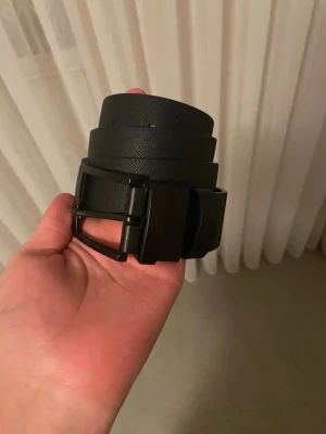 Svart bälte med klassiskt spänne - Snyggt svart bälte i skinnimitation med ett stilrent, matt svart spänne. Perfekt för att lyfta vilken outfit som helst och passar till både jeans och kostymbyxor. Enkel design utan mönster för en clean look.