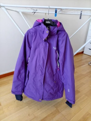 Lila vindjacka från North Bend - Snygg lila vindjacka från North Bend med huva och dragkedja. Jackan har rosa innerfoder, praktiska fickor och justerbar huva med snören. Perfekt för blåsiga dagar och har en sportig look. Materialet är slitstarkt syntet som står emot vind. De ändå ä att dragkedjan ha tappat bandet men går å sätta ditt ett nytt å färgerna ha tappat lite färg i