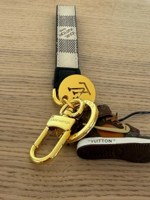  Vuitton nyckelring med sneaker - Unik nyckelring från  Vuitton med guldfärgade metalldetaljer, beige och brunt band samt en miniatyrsneaker i brunt och orange med LV-monogram. Perfekt accessoar för dig som gillar streetstyle och lyxiga detaljer.