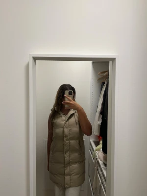 Zara lång dunväst – beige – strl S 🤍 - Snygg beige lång dunväst med hög krage och dragkedja framtill. Västen har två sidofickor och quiltad design som ger en trendig look. Perfekt att slänga över en hoodie eller tröja för extra värme. Passar till många olika stilar.