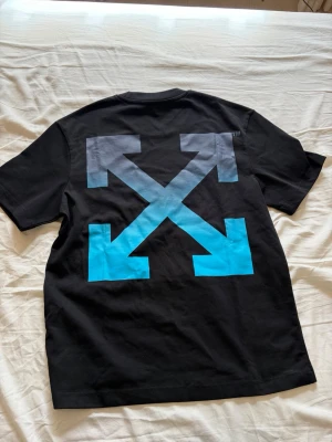 Svart Off-White t-shirt med blå tryck - Säljer en svart t-shirt från Off-White med stort blått och grått pilar-tryck på ryggen och Off-White-logga i blått framtill. Klassisk passform och rund hals. Perfekt för dig som gillar streetwear och vill sticka ut med en ikonisk design.