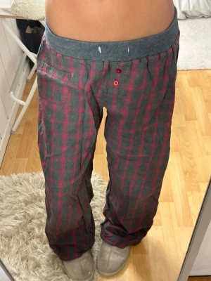 Rutiga pyjamasbyxor i grått och rött - Mysiga pyjamasbyxor med grå bas och rött rutmönster. Byxorna har en bred, mjuk midjeresår i mörkgrått och knappdetaljer framtill. Perfekta för chillkvällar hemma. Materialet känns mjukt och bekvämt mot huden.