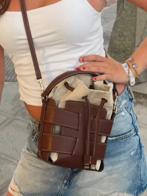 Brun axelväska i läderlook - Snygg brun axelväska i läderlook med justerbar rem och coolt flätat mönster. Väskan har en beige tygpåse inuti som stängs med dragsko. Perfekt för dig som vill ha en trendig och unik accessoar till din outfit.