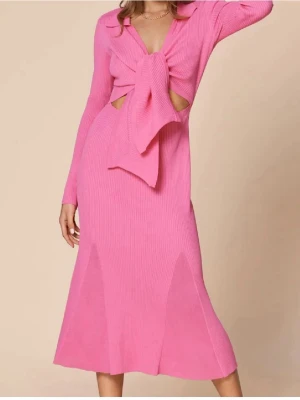 Rosa ribbad långklänning med knyt - Trendig rosa långklänning i ribbat material med lång ärm och stor knytning framtill. Klänningen har krage, v-ringning och snygga cut-outs vid midjan för en modern look. Perfekt för dig som vill sticka ut med en färgstark och unik stil.