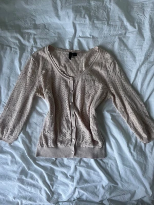 Ljusbeige virkad kofta med knappar - Supersöt ljusbeige kofta med virkade detaljer och knappar framtill. Koftan har trekvartsärmar, rund halsringning och ribbade muddar. Perfekt att slänga över en topp för en chill look.