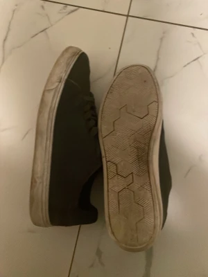 Svarta låga sneakers med vit sula - Säljer ett par svarta sneakers med klassisk låg profil och vit sula. Skorna har snörning och en enkel, stilren design som passar till det mesta. Yttermaterialet ser ut att vara textil eller syntet och sulan har ett hexagonmönster undertill.