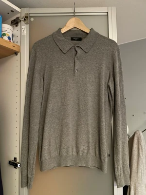 Grå långärmad polotröja  - Snygg grå långärmad polotröja med krage och tre knappar framtill. Tröjan är i mjukt stickat material och har ribbade muddar vid ärmslut och nederkant. Perfekt för en stilren och enkel look. Den är från Matinique.  
