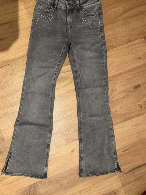 Grå utsvängda jeans med nitar - Säljer ett par grå jeans från LINDEX med utsvängda ben och slitsar nertill. Jeansen har dekorativa små nitar i silver och pastellfärger vid fickorna fram. Klassisk femficksmodell och skön stretchig denim. Perfekta för en trendig och avslappnad look.