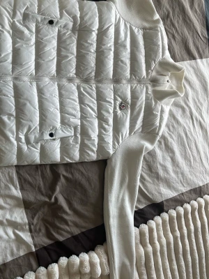 Vit cardigan - Snygg vit cardigan med quiltad, vadderad front och stickade ärmar. Jackan har två framfickor med knappstängning, dragkedja framtill och logga på bröstet samt broderad logga på ärmen. Perfekt för lager-på-lager och streetstyle. Använd endast en gång säljer eftersom jag ej använder dessa typer av kläder längre. Priset går att diakuteras vid snabb affär