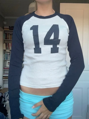Vit och marinblå långärmad topp - Sportig långärmad topp med marinblå ärmar och vit kropp. Stort tryck med siffran 14 framtill. Ribbad struktur och tight passform, perfekt för en avslappnad och trendig look.