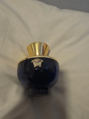 Versace blå parfymflaska guld - Elegant parfymflaska från Versace i djupblått glas med guldfärgade detaljer och Versaces ikoniska Medusa-logga framtill. Flaskan har en rundad form och en lyxig känsla med sin guldfärgade topp och spridare.