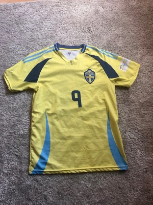 Svensk gul fotbollströja nr 9 - Gul fotbollströja med blå detaljer och nummer 9 på framsidan. Tröjan har Sveriges landslagsemblem på bröstet, V-ringad krage och blå ränder på axlarna. Perfekt för dig som vill stötta Sverige på match eller träning.